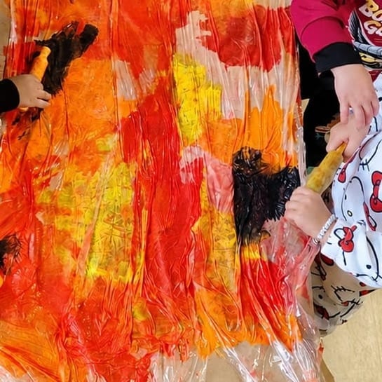Manos de varios niños pasando rodillos de pintura naranja, roja y amarilla sobre un plástico extendido en la mesa, en una propuesta de pintura colectiva