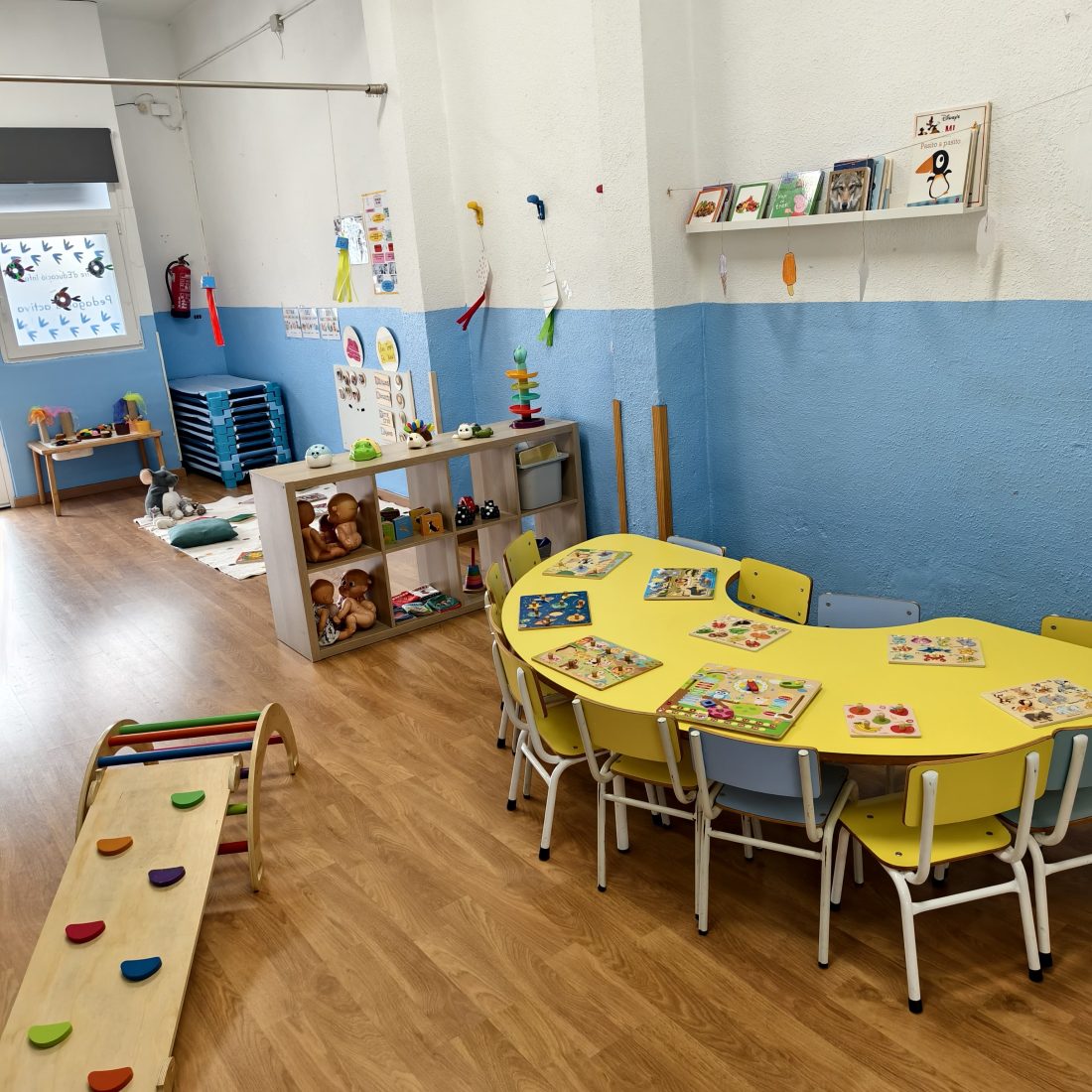 Aula de Ratolins vacía, con la mesa amarilla preparada con encajables, la estantería de material al alcance y la rampa de psicomotricidad