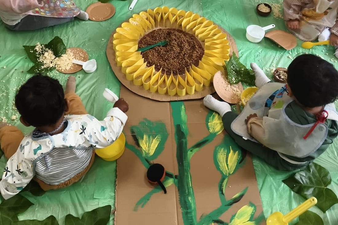 Dos bebés de espaldas, sentados en el suelo alrededor de un girasol grande de cartón relleno de sémola, con cuencos, cucharas y hojas repartidos alrededor