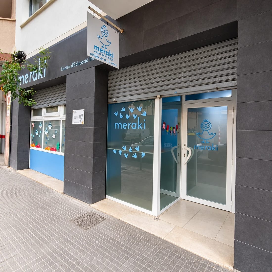 Fachada de Meraki en el carrer de Niceto Alcalá Zamora: rótulo colgante con el pollito de la marca, la entrada acristalada y la ventana del aula decorada con dibujos de los niños