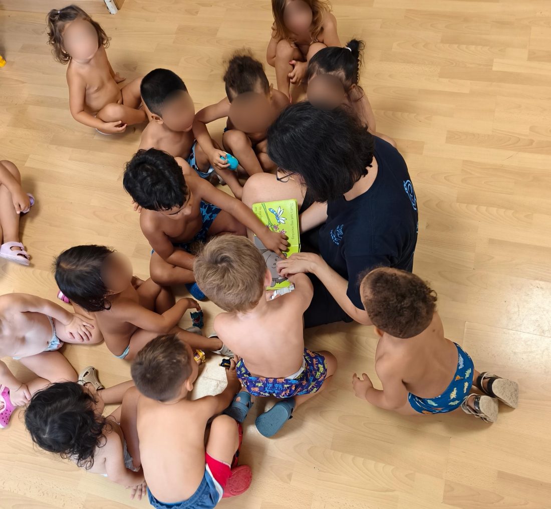 Montserrat Losada, de espaldas, leyendo un cuento a un grupo de niños sentados en círculo en el suelo del aula, con las caras difuminadas