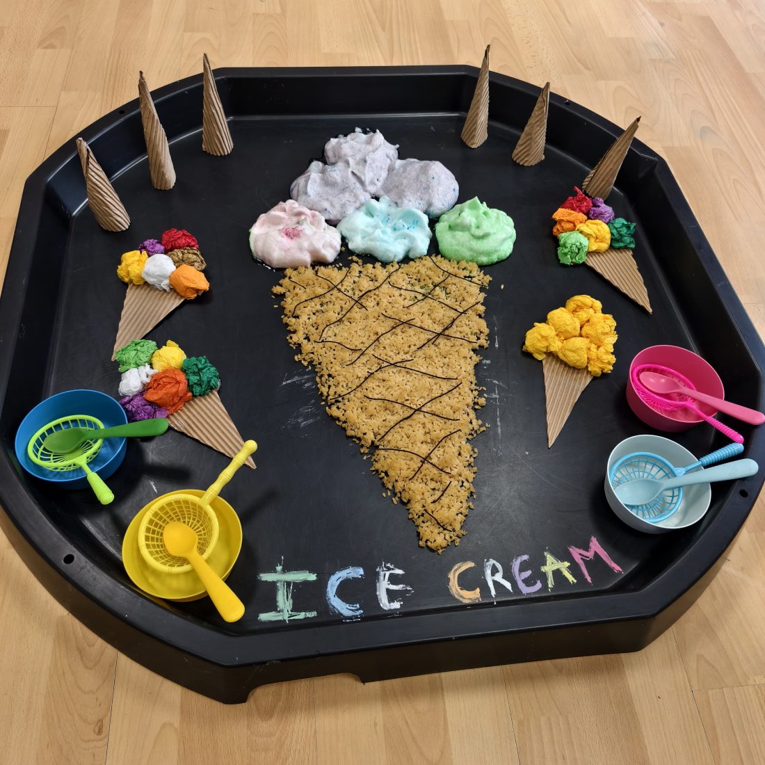 Bandeja de experimentación del aula de los pequeños preparada como una heladería, con espuma de colores, arroz teñido, conos de cartón y cuencos con cucharas