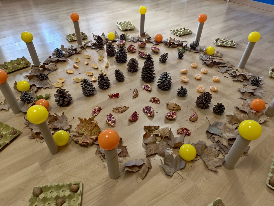 Un círculo de piñas, granadas, nueces y hojas secas alrededor de tubos de cartón rematados con bolas de colores, para una propuesta de otoño