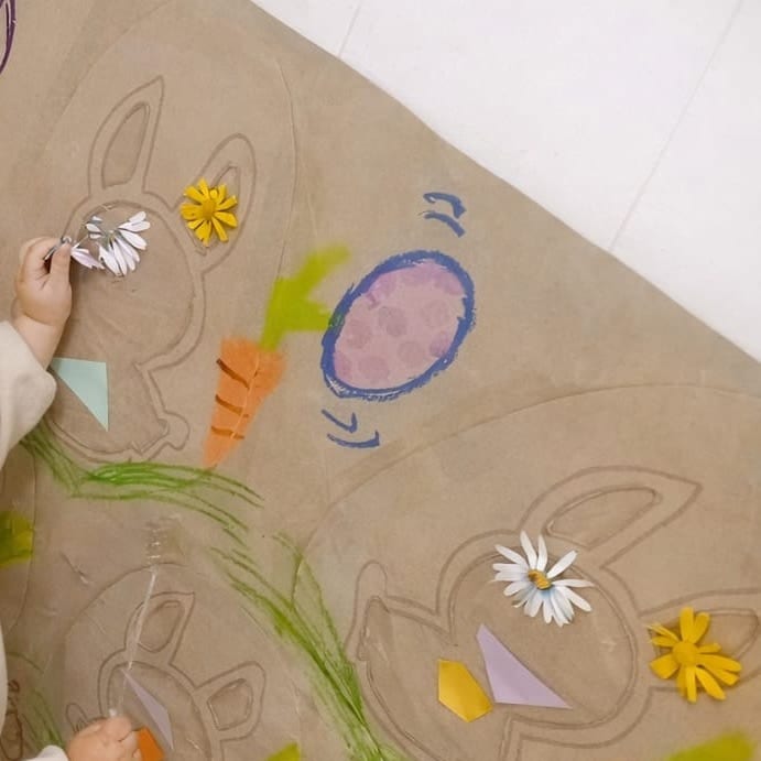 La mano de un niño pegando una margarita de papel en un mural de conejos dibujado sobre papel kraft, colocado a su altura